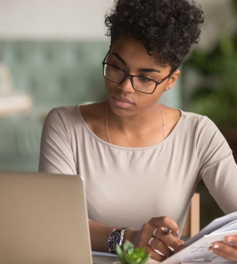 A focused woman with glasses reading a book beside her laptop